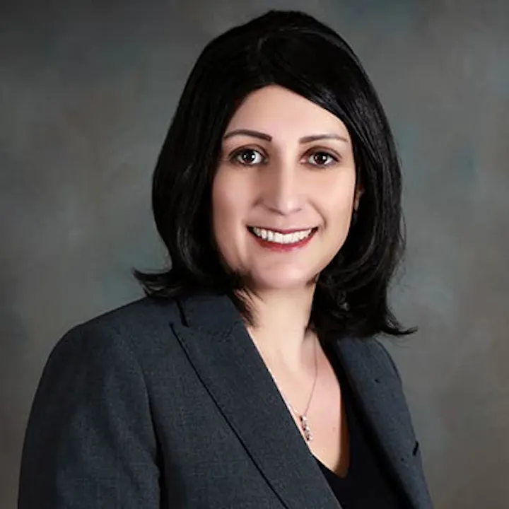 A professional headshot of a woman with dark hair, wearing a blazer, smiling against a blurred background.