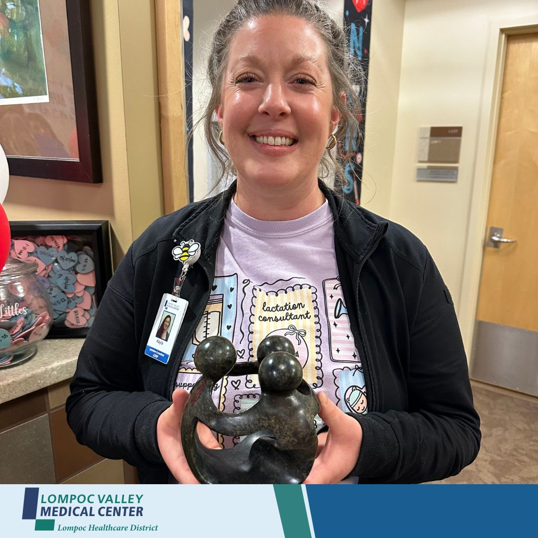 A woman holds a sculpture while smiling, wearing a shirt labeled "lactation consultant," in a medical center setting.