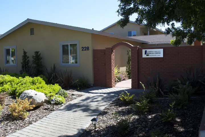 The image shows the exterior of Lompoc Valley Medical Center, featuring a pathway and a welcoming entrance with greenery.