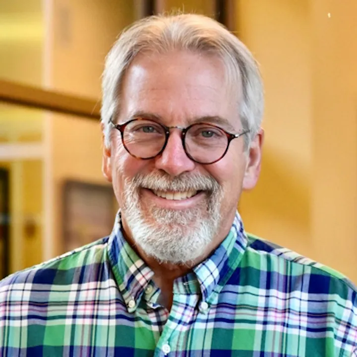 An older man with glasses and a friendly smile, wearing a colorful plaid shirt, is standing in an indoor setting.