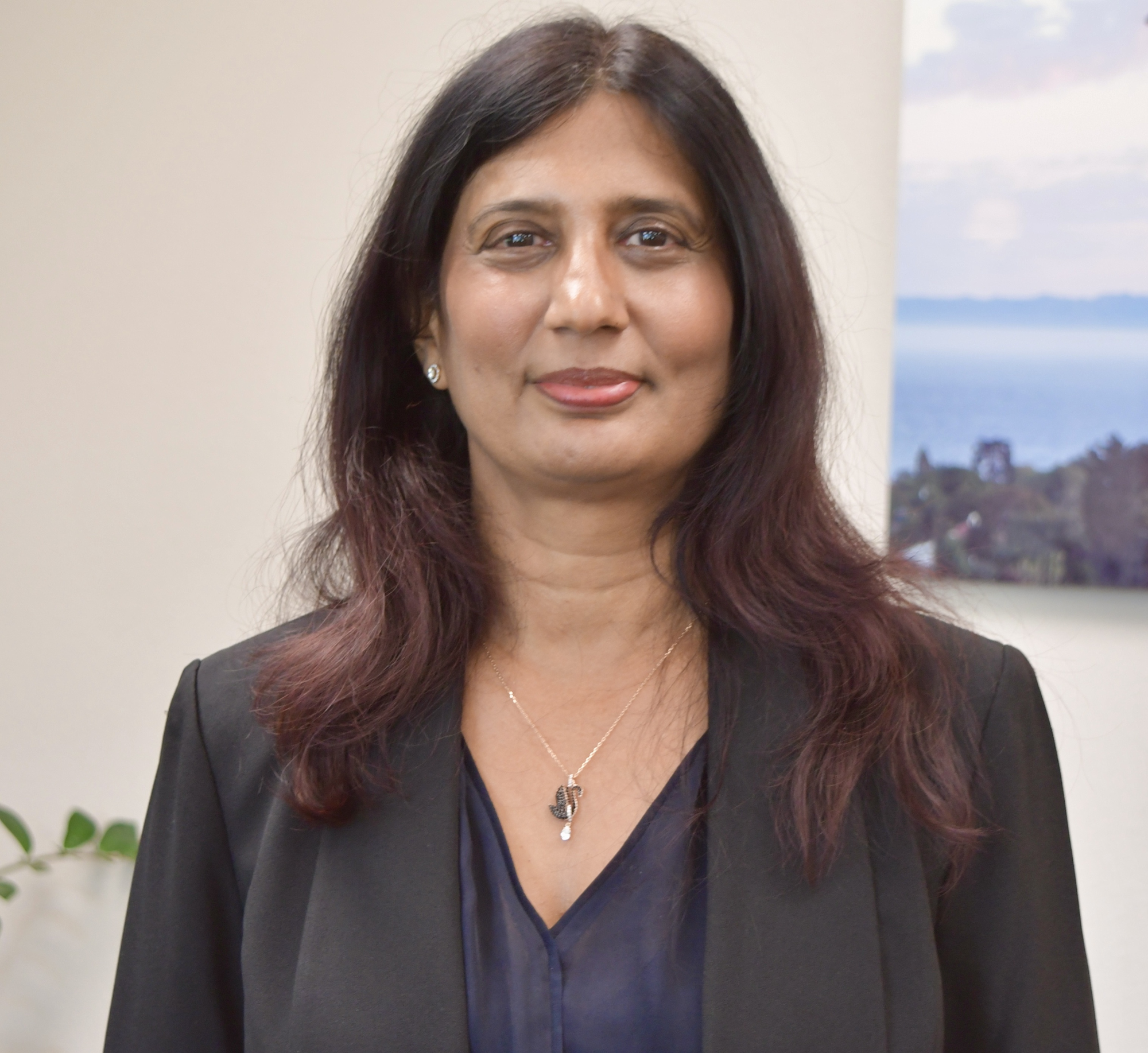 The image shows a woman with long hair, wearing a dark blazer and a necklace, standing in an office setting.