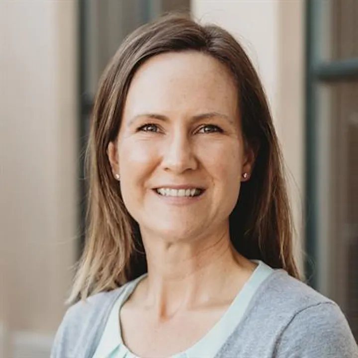 The image features a woman with long hair, smiling warmly, wearing a gray sweater and a light blue top against a neutral background.