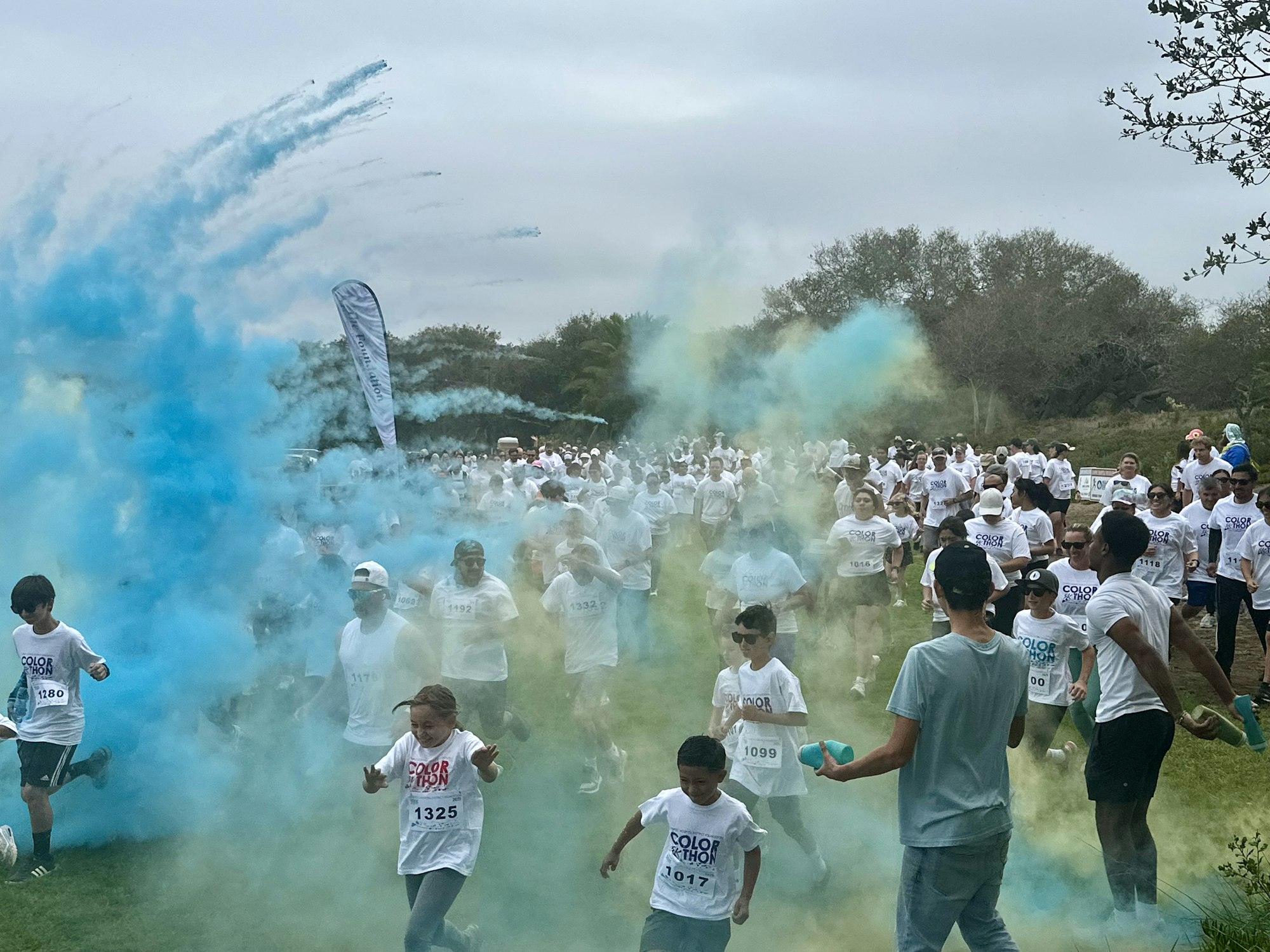 A lively color run event with participants in white shirts surrounded by vibrant blue smoke and playful energy.