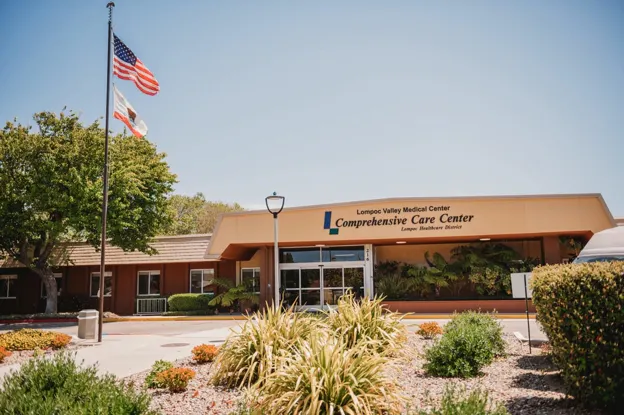 The image shows the exterior of the Lompoc Valley Medical Center Comprehensive Care Center, surrounded by greenery and flags.