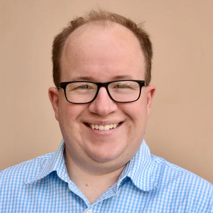 A smiling person with glasses, wearing a blue checkered shirt, poses in front of a neutral background.