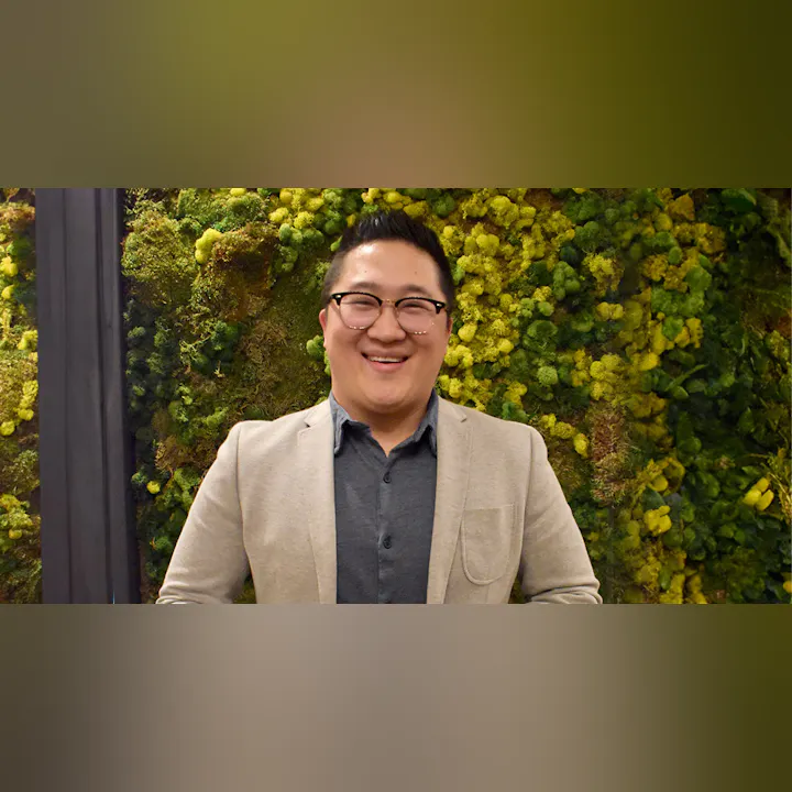 A smiling person in a light blazer stands against a vibrant green moss wall, creating a lively and inviting atmosphere.