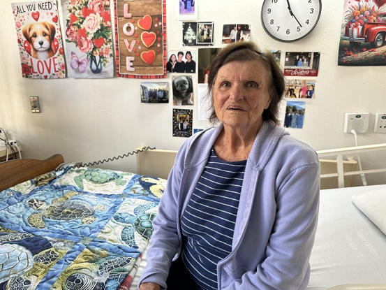An elderly woman in a light sweater sits on a bed with a patterned quilt, surrounded by decorative photos and a clock.