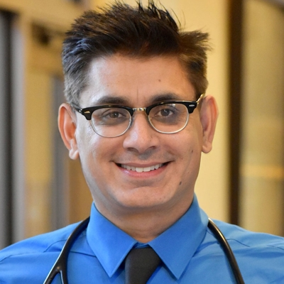 The image features a smiling man in a blue shirt and tie wearing glasses, with a stethoscope around his neck.