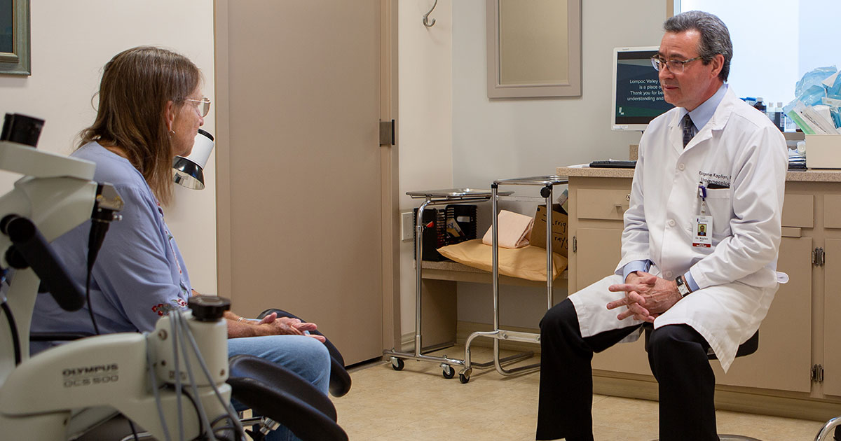 A doctor consults with a patient in a medical office setting, discussing her health concerns. Medical equipment is visible.