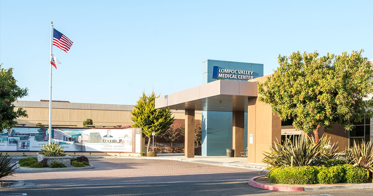 Entrance of Lompoc Valley Medical Center with American and Californian flags, surrounded by greenery and a clear sky.
