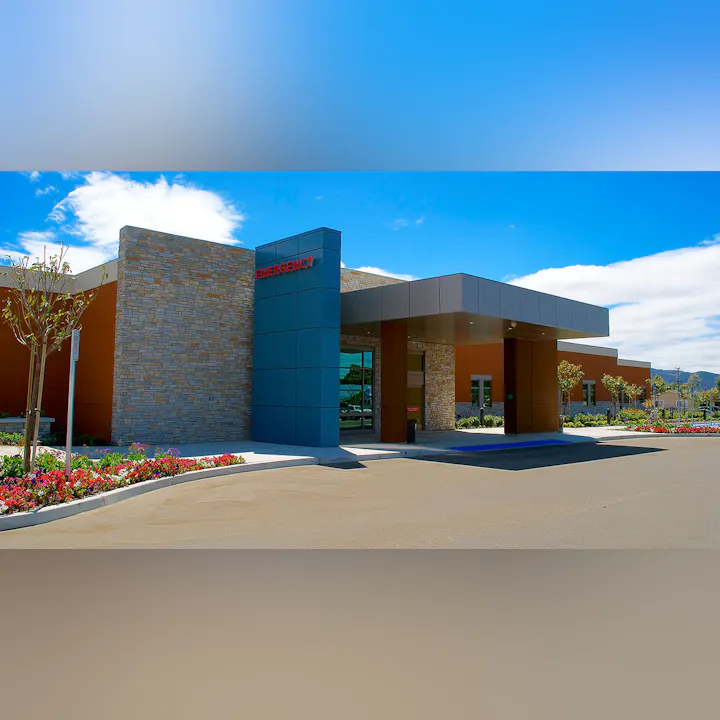 The image shows a modern emergency room entrance with a clear blue sky and landscaped gardens.