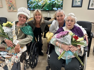Two women in wheelchairs, dressed as queens with bouquets, pose with two smiling caregivers in a cheerful setting.