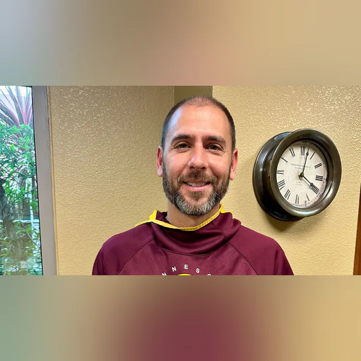 A smiling man with a beard is wearing a maroon hoodie. There's a clock on the wall behind him and plants visible outside.