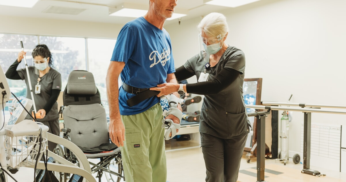 A patient is assisted by a therapist in a rehabilitation setting, focusing on walking with supportive equipment.