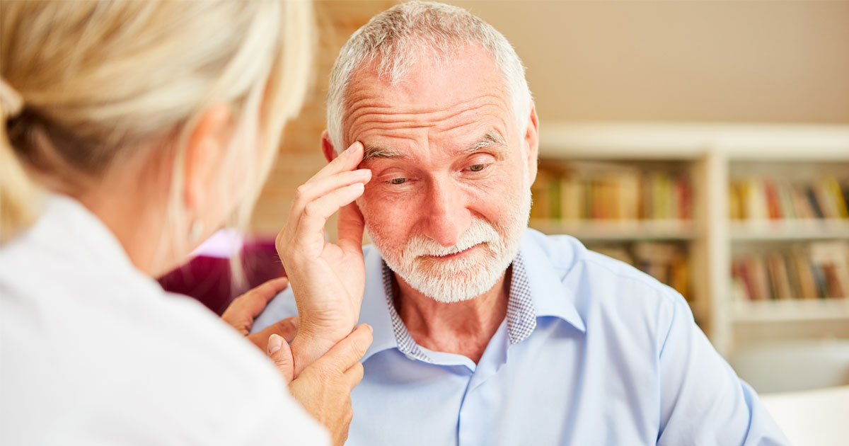 A female caregiver gently interacts with an elderly man, who appears contemplative, in a cozy indoor setting with bookshelves.