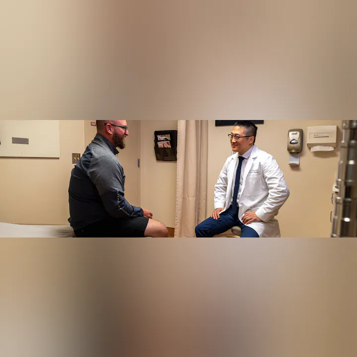 A doctor in a white coat chats with a patient in a medical office, both appearing engaged in conversation.