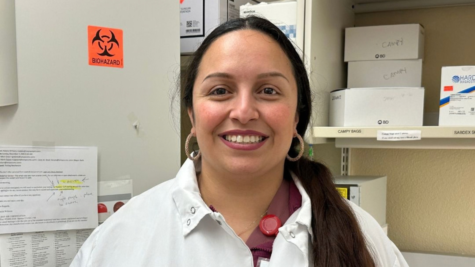 A person in a lab coat smiles in a lab setting, beside a biohazard sign and shelves with labeled boxes.