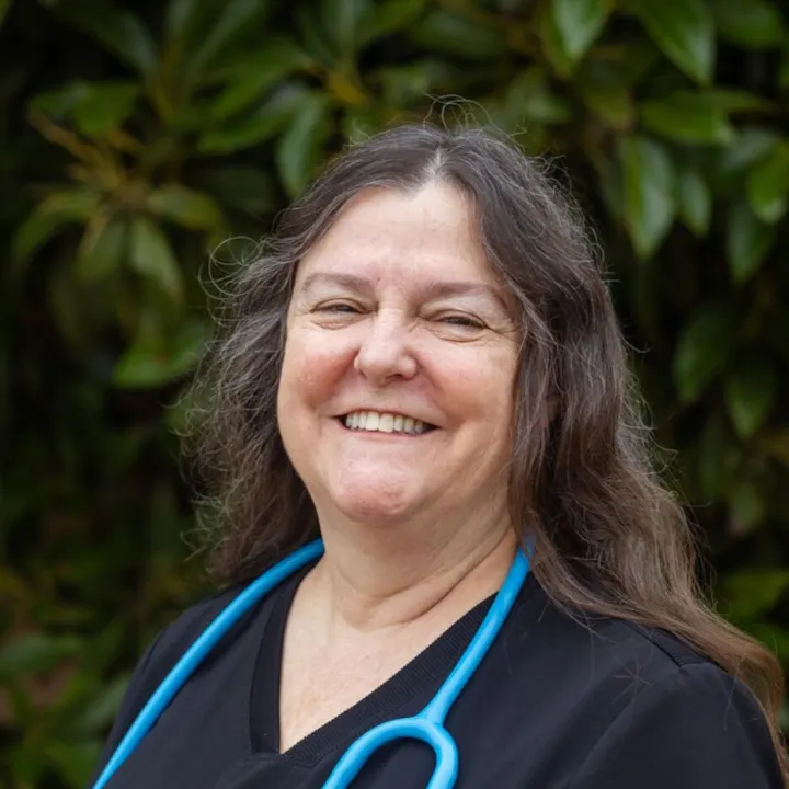 A smiling person with long hair wears scrubs and a stethoscope, with greenery in the background.
