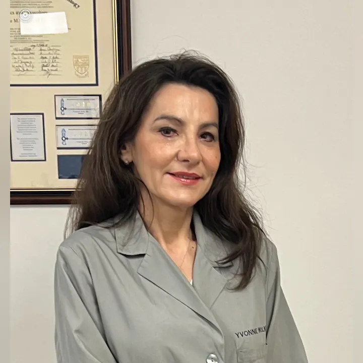 A woman in a gray coat stands smiling in front of a framed document and certificates on a wall.