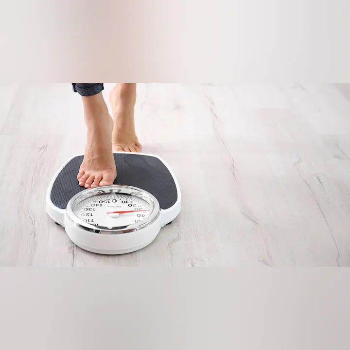 A foot is on a weighing scale, highlighting the act of weighing oneself, with a wooden floor backdrop.