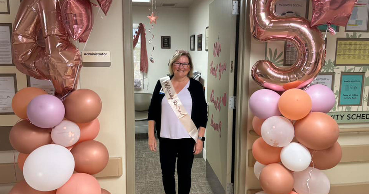 A woman in a black top with a sash celebrates an event, surrounded by decorative balloons and a large number "5."