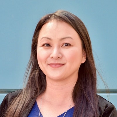 A woman with long hair is smiling in front of a blue background, wearing a blue top and a black jacket.