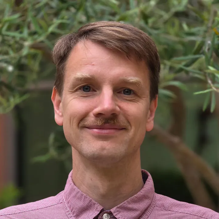 A smiling man with a mustache is posed in front of a green background with foliage.