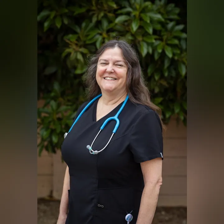 A smiling woman in a black medical uniform with a blue stethoscope stands outdoors among green foliage.