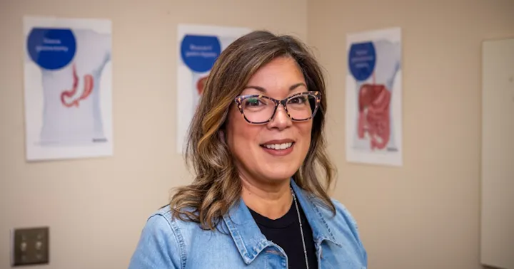 A smiling woman in a denim jacket stands in a medical setting, with anatomical posters on the wall behind her.