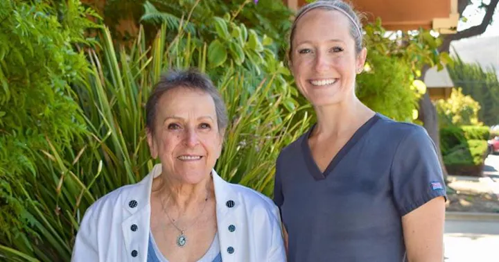 Two women are smiling together outdoors, surrounded by greenery, with one dressed in a white jacket and the other in scrubs.