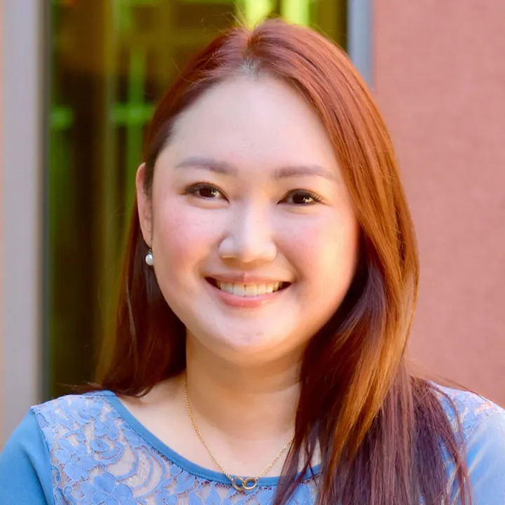 A smiling person with long reddish-brown hair, wearing a blue top with lace details, in front of a colored backdrop.