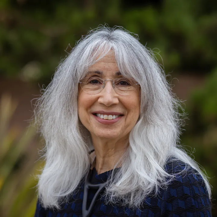A woman with long gray hair, glasses, and a friendly smile is standing outdoors against a blurred green background.