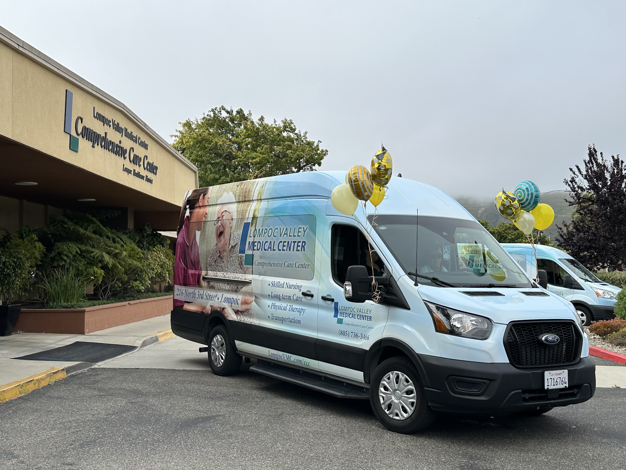 The image shows a medical center building with a van parked outside, decorated with balloons, promoting the Lompoc Valley Medical Center.