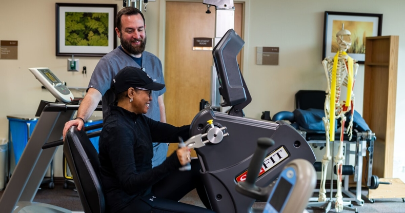 A woman exercises on a fitness machine with guidance from a trainer in a gym-like setting.