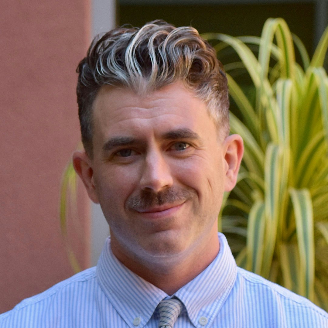 A person with styled hair and a mustache smiles, wearing a collared shirt with a striped pattern, against a leafy background.