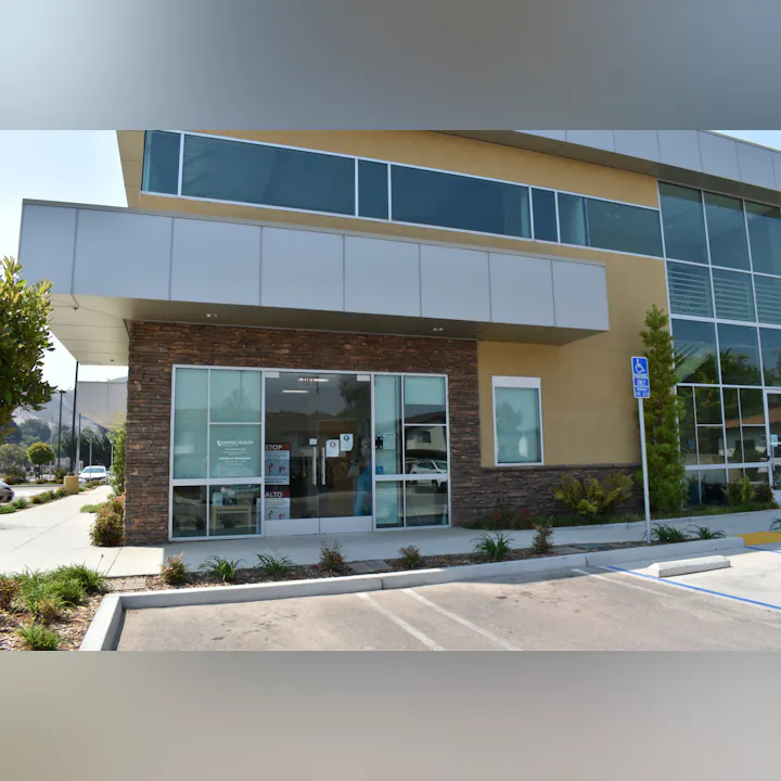 The image shows a modern health facility facade with large glass windows and a handicapped parking sign.