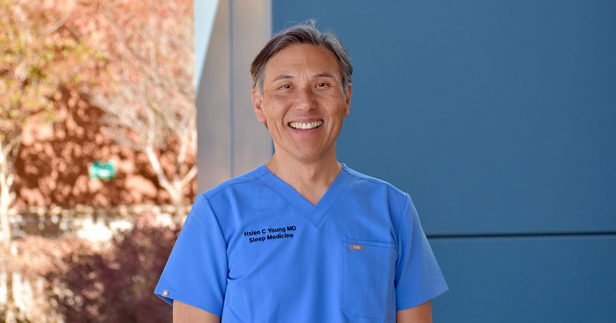A smiling medical professional in blue scrubs stands outdoors, with trees and a modern building in the background.