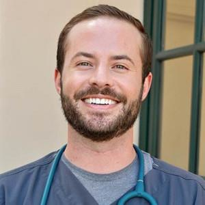A smiling person wearing medical scrubs and a stethoscope, posed outside a building with windows.