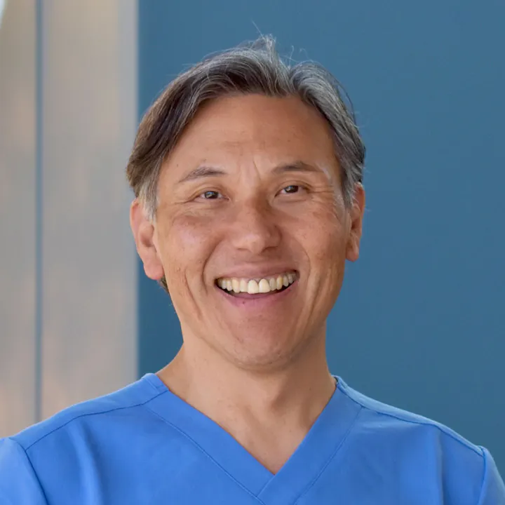 A smiling person in a blue medical scrub stands in front of a blue wall, exuding a friendly and approachable vibe.