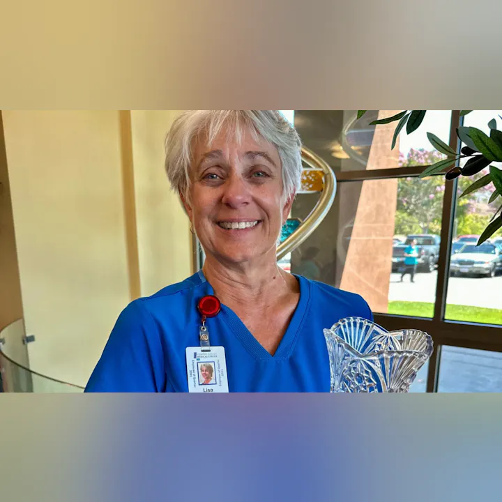 A smiling woman in scrubs holds a crystal award, standing in a well-lit area with greenery and cars visible outside.