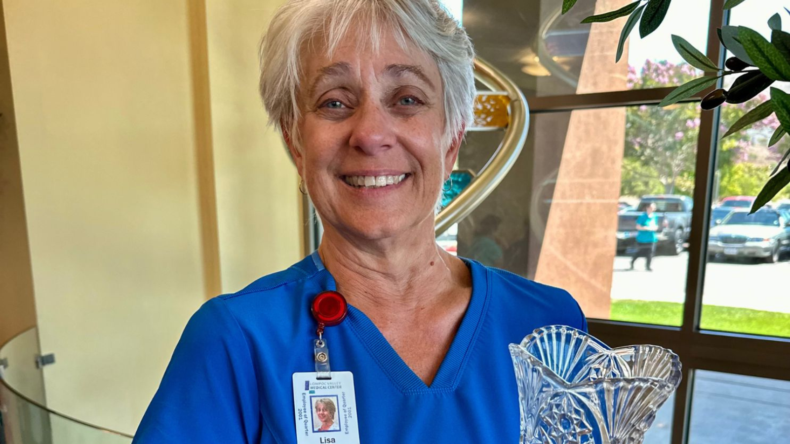 A smiling woman in scrubs holds a crystal award, standing in a well-lit area with greenery and cars visible outside.