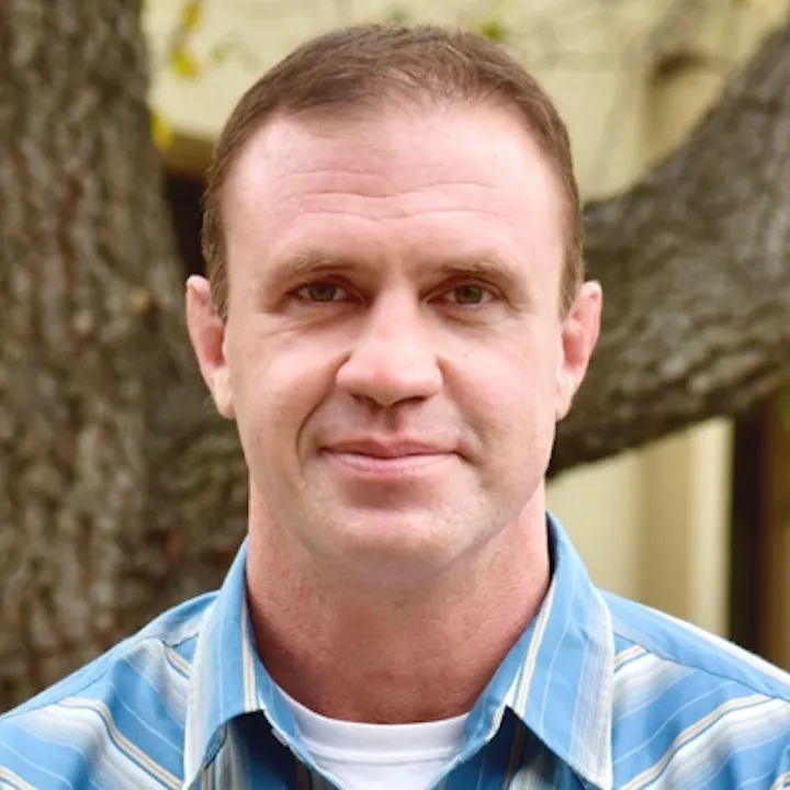 A man with short hair is smiling, wearing a striped blue shirt, with a tree in the background.