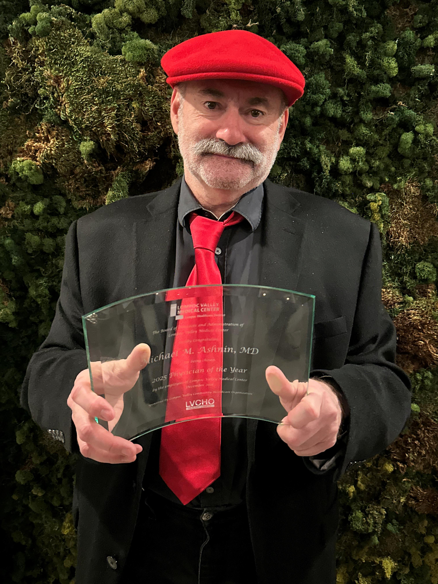 A man wearing a red hat and tie holds an award recognizing him as "2025 Physician of the Year" from a medical center.