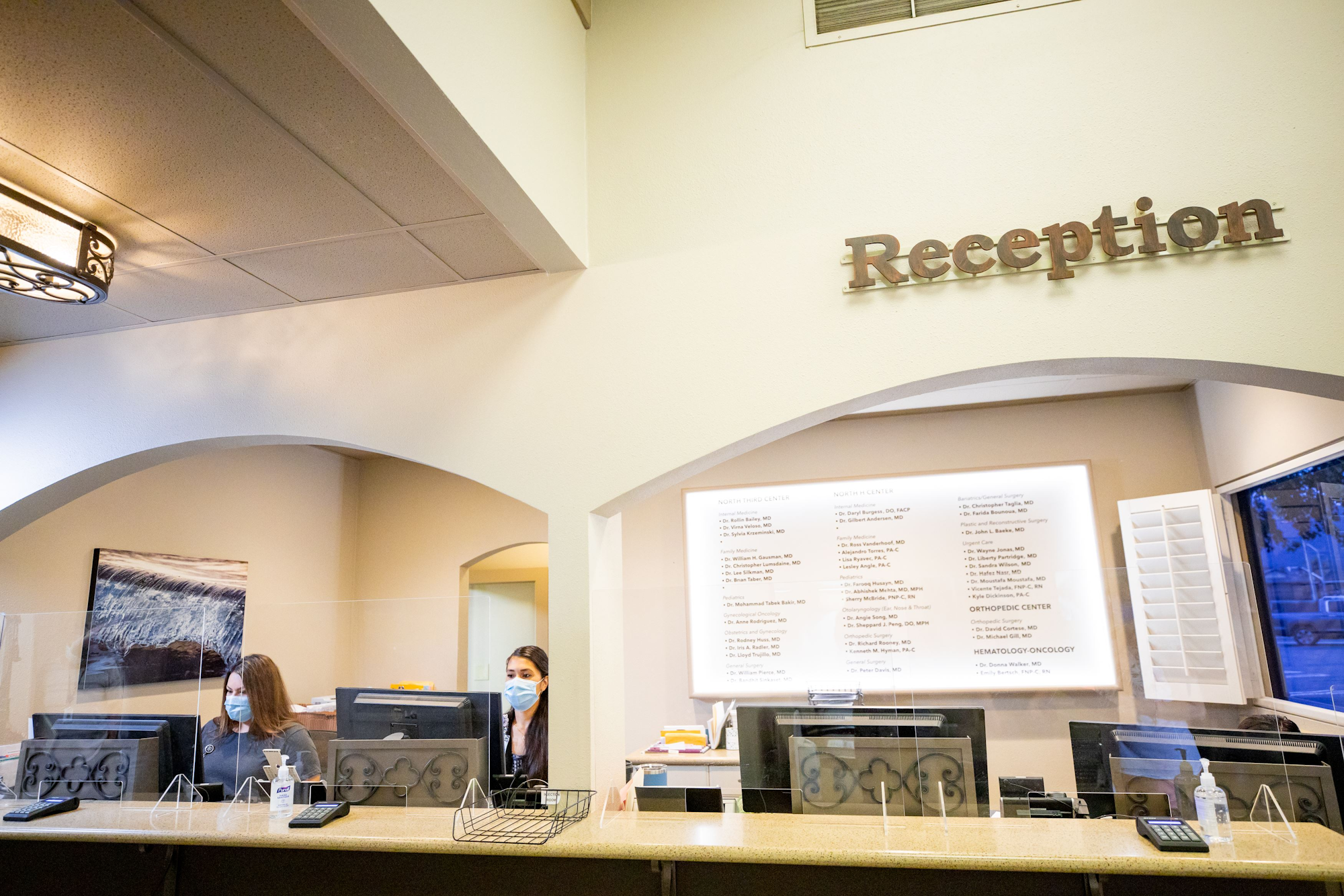 The image shows a reception area with staff wearing masks, computers, and a list of services on the wall.
