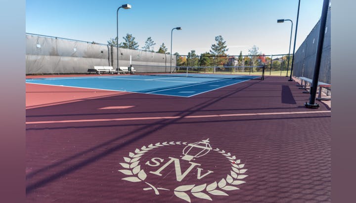 An empty tennis court with a distinctive logo on the foreground surface and benches alongside.