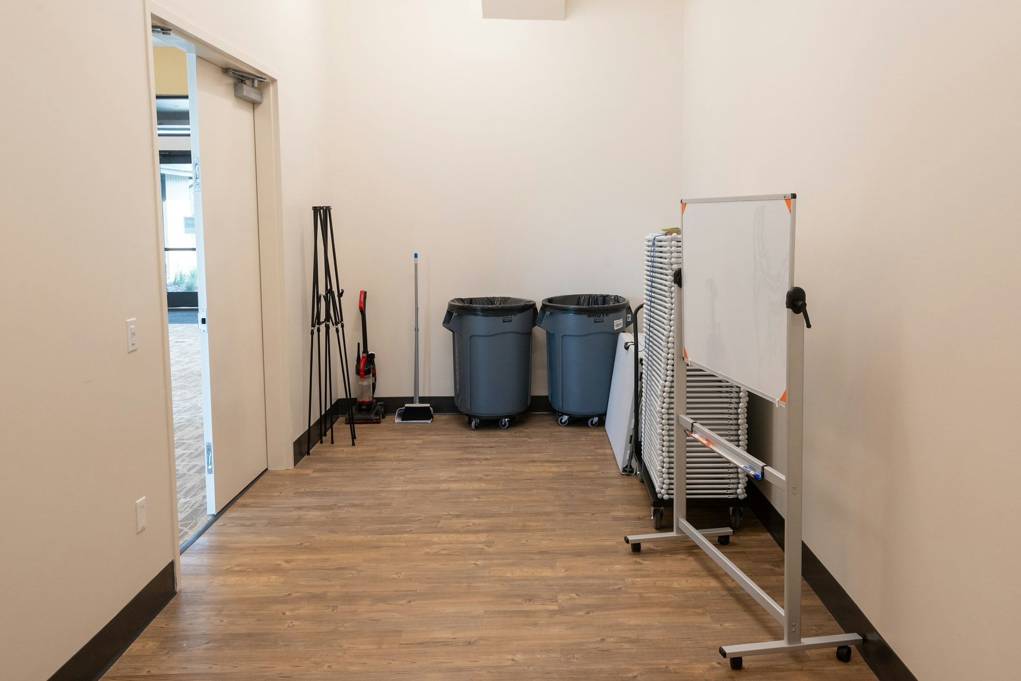 A hallway with cleaning supplies, trash cans, and stacked chairs next to a whiteboard.
