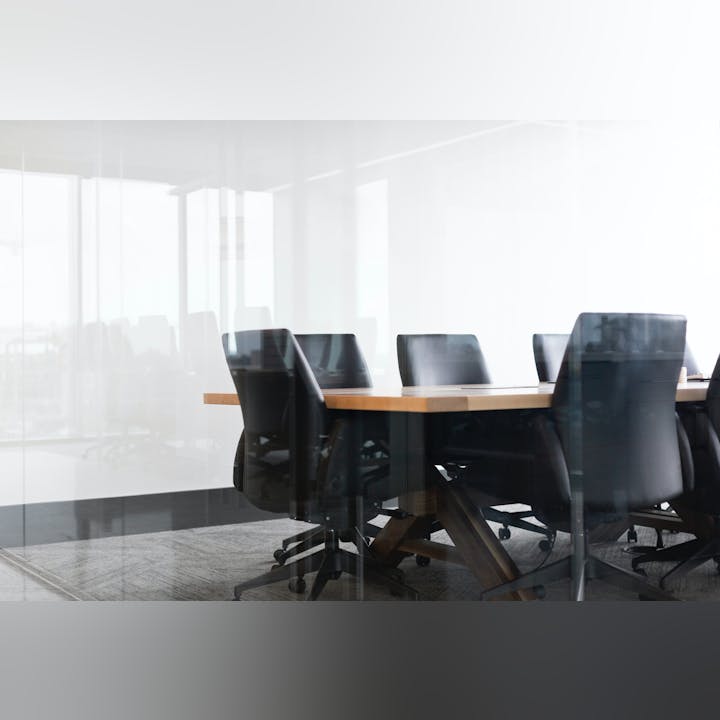 An empty, modern conference room with a wooden table and black office chairs, seen through a glass wall.