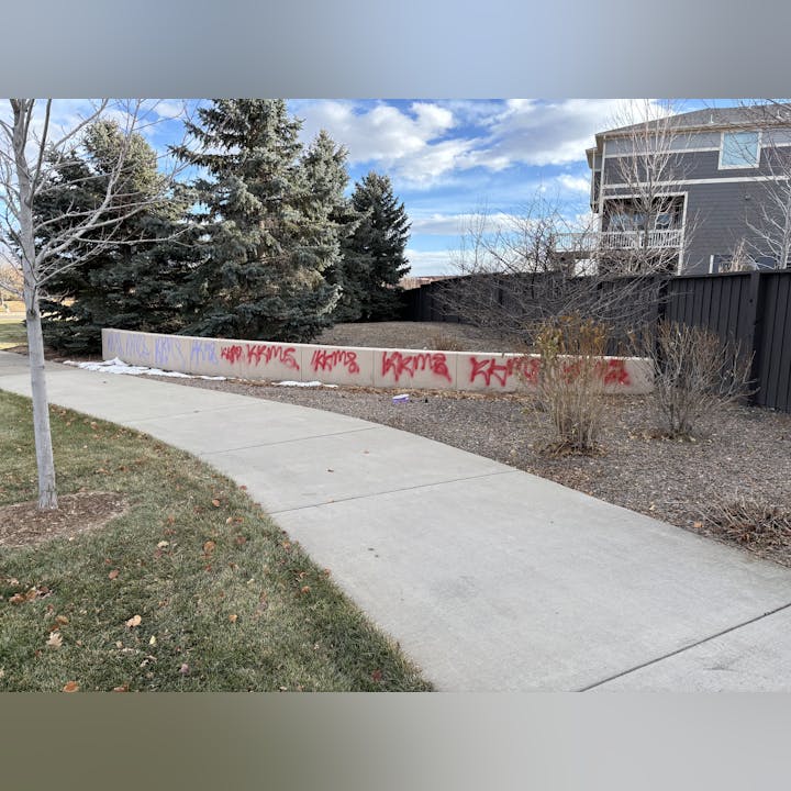 Concrete wall with graffiti near a sidewalk, trees, and a residential building in the background.