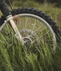 A close-up of a motorcycle wheel partially obscured by tall grass, showcasing its tire and brake components.