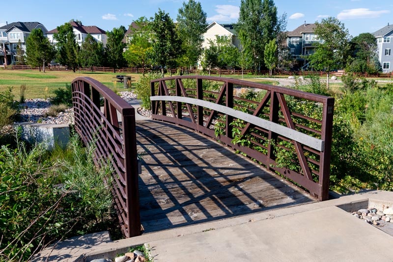 Wooden footbridge with metal railings over a small creek in a suburban area, with houses in the background.